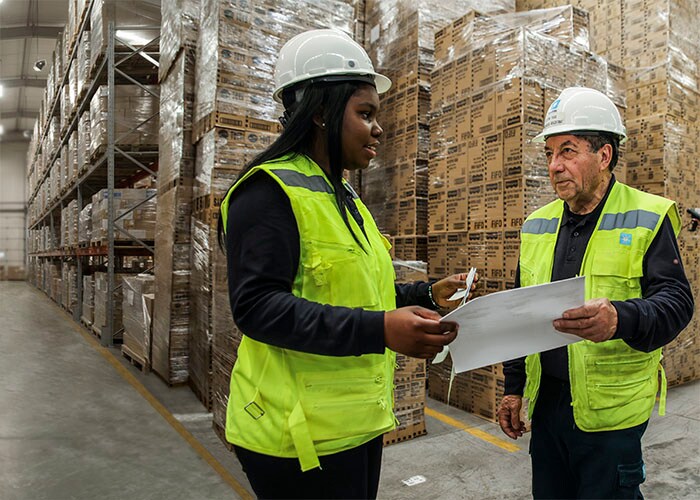 Two workers wearing high‑visibility vests and hard hats review documents together inside a warehouse with stacked pallets and shelving.