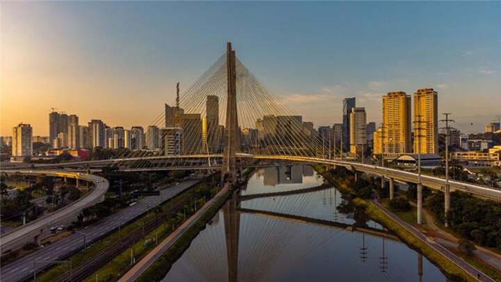 Aerial view of the Octávio Frias de Oliveira Bridge and surrounding São Paulo skyline at sunset, with the bridge reflecting on the river below.