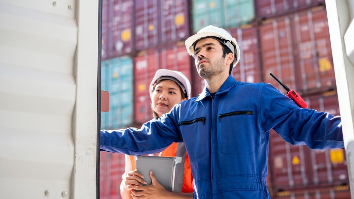 Two workers wearing safety helmets and uniforms stand in front of a shipping container.