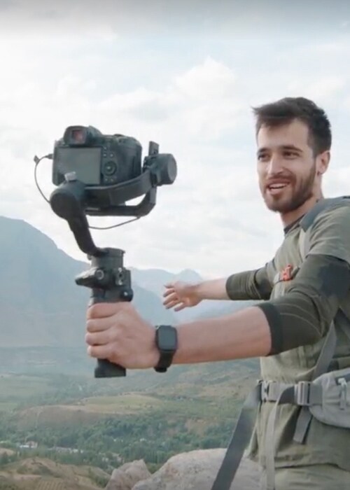 Travel vlogger filming with a DSLR camera on a stabilizer while hiking in the mountains with a backpack and climbing rope.