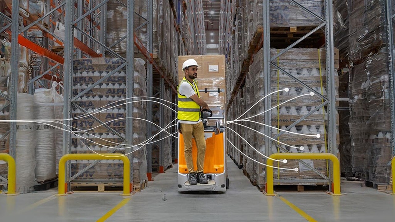 A man operating a forklift inside a warehouse, surrounded by shelves of stored goods.