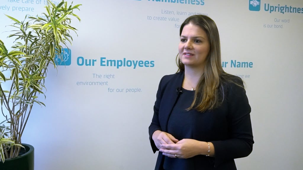 A professional woman in a dark blazer giving a presentation in front of a company values wall, with words like 'Humbleness,' 'Uprightness,' and 'Our Employees' visible in the background.