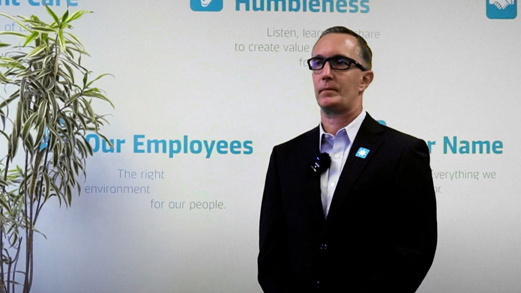 A professional man in a dark suit and glasses delivering a speech in front of a company values wall, with words like 'Humbleness,' 'Our Employees,' and 'Constant Care' visible in the background.