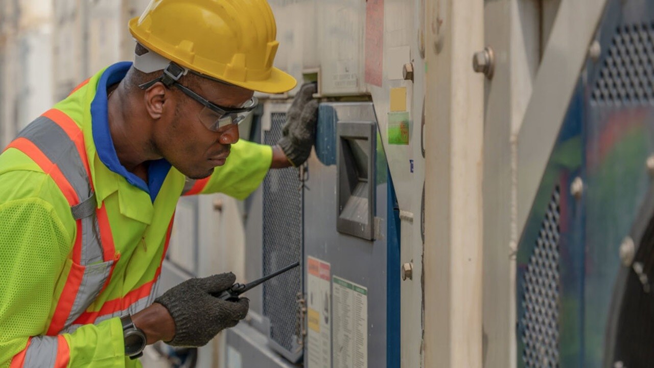 A man wearing a hard hat and safety vest inspects a machine for maintenance or safety checks.
