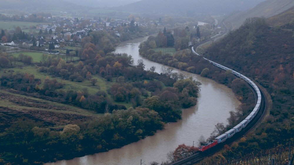 A Maersk freight train winding alongside a river through a lush, rural landscape with small villages and hills in the background.