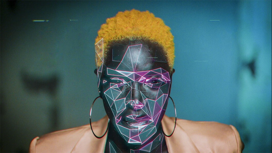 A black woman with short, yellow hair and wearing a peach-coloured suit jacket stares into the camera