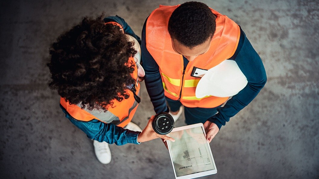 Two people wearing orange hi-vis vests over their clothes look at a tablet showing a digitised stock list