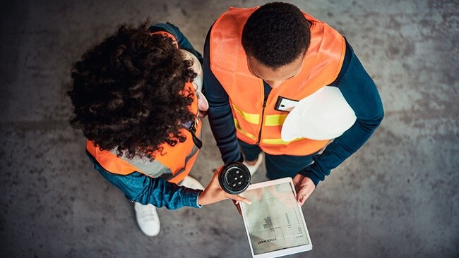 Two people wearing orange hi-vis vests over their clothes look at a tablet showing a digitised stock list