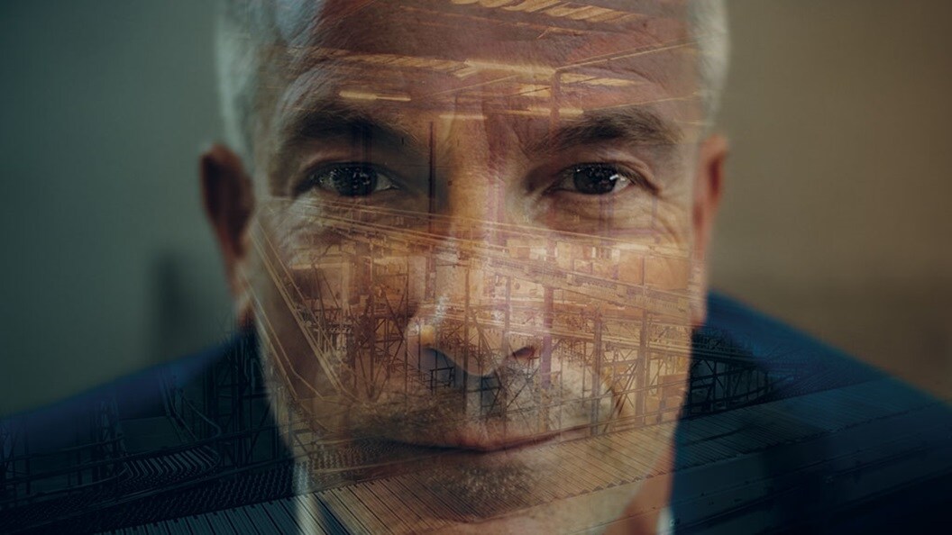 A man with white hair looks straight into the camera. Overlaid is an image of warehouse conveyor belts carrying cardboard boxes
