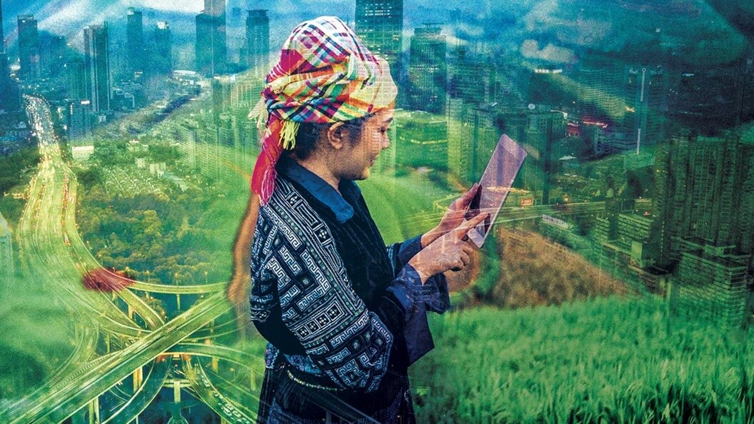 A person wearing a colourful headscarf and quilted jacket is standing in a paddy field and looking at their tablet computer.