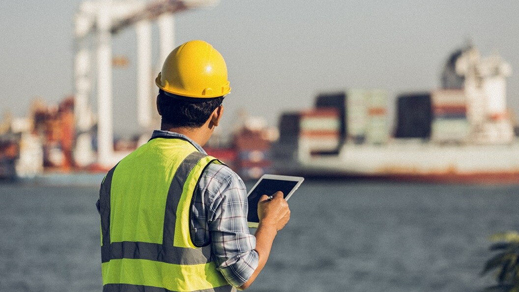 A person in a hard hat holds a tablet and looks out at a shipping terminal, symbolising digitalisation in shipping and logistics