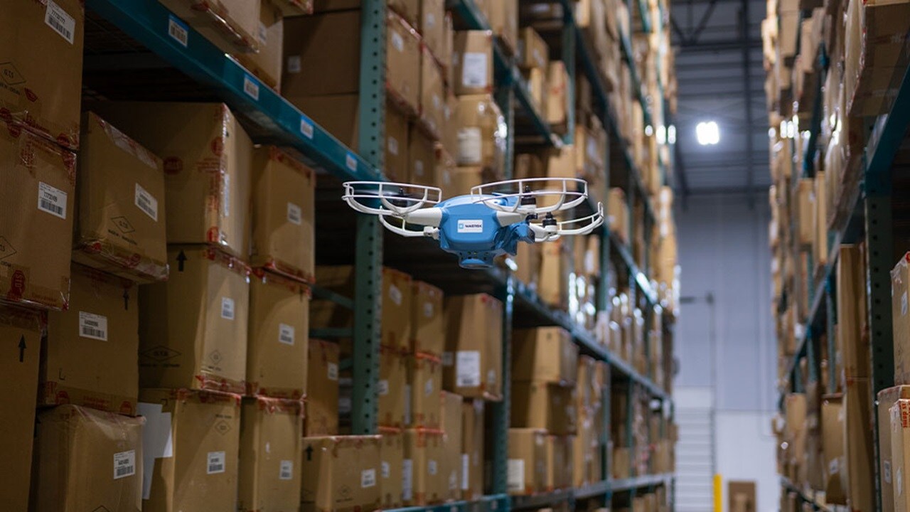 A blue drone hovers in a large warehouse filled with storage racks.