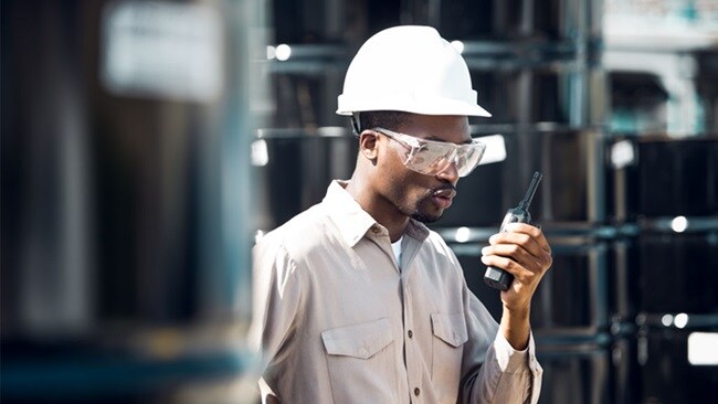 a man wearing a white hat and overalls talks into a walkie talkie while walking past large industrial tanks