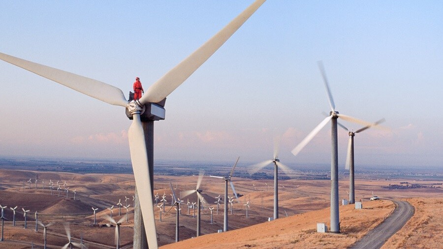 A worker in red safety gear sits atop a wind turbine, overseeing a vast wind farm on rolling hills under a clear sky.