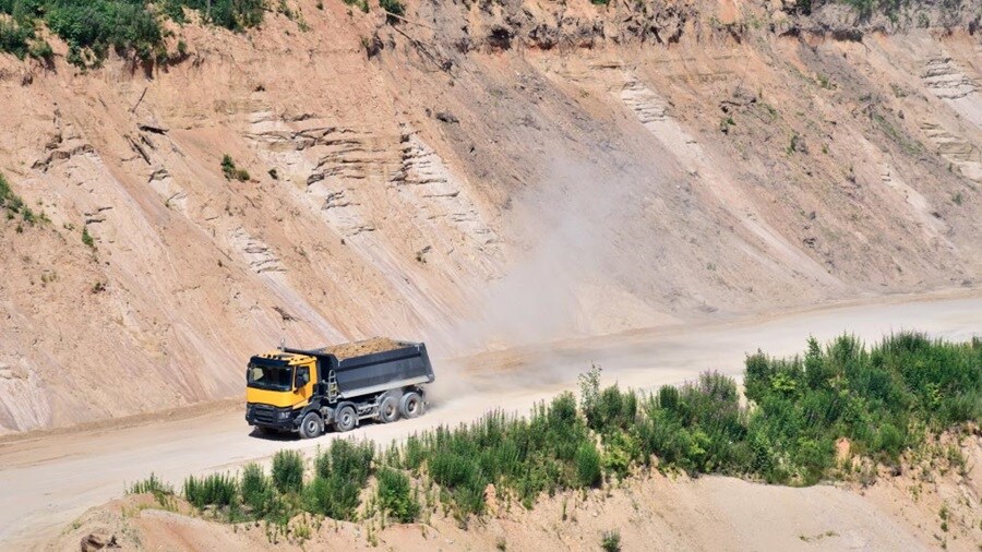 A yellow dump truck transports soil along a dusty road in a rugged quarry, surrounded by steep rock formations and greenery.