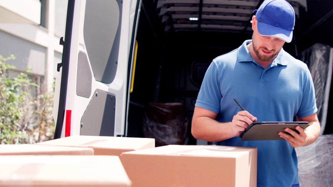 A man wearing a blue shirt stands in front of a van, holding a tablet in his hands.