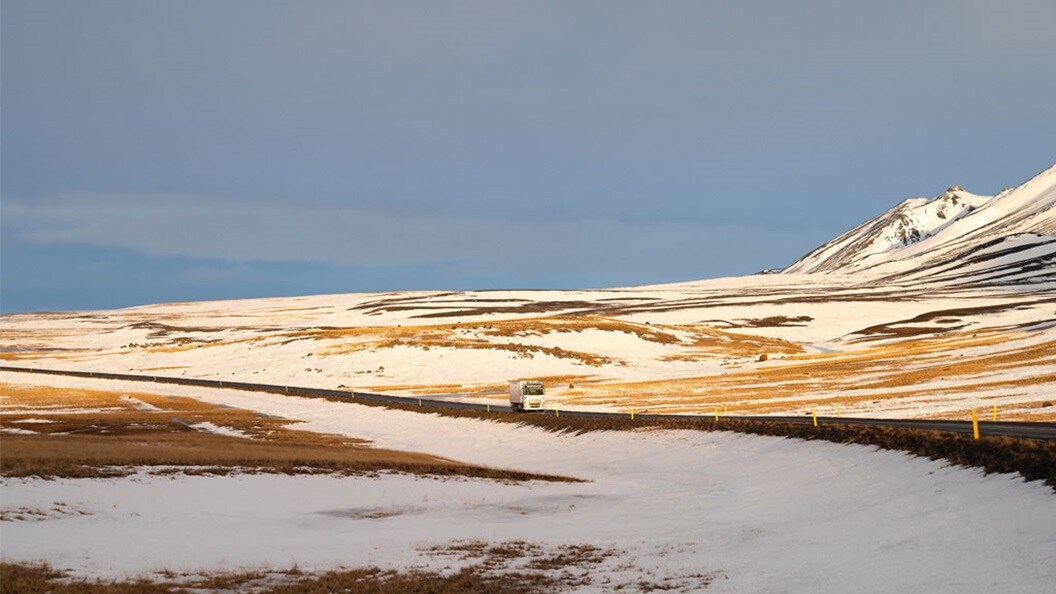 A white truck driving on a long street in between white snowy mountain edge.