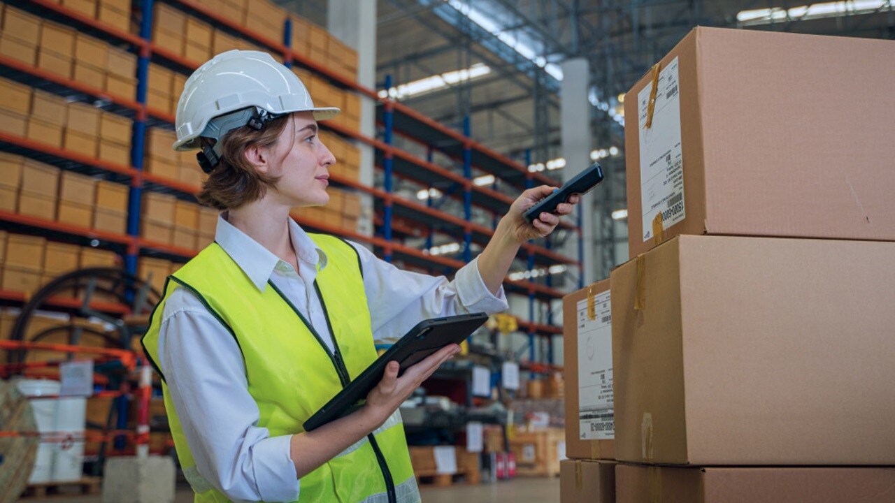 A person wearing a white hard hat and high-visibility vest is holding a barcode scanner and a clipboard, standing in front of cardboard boxes in a warehouse.