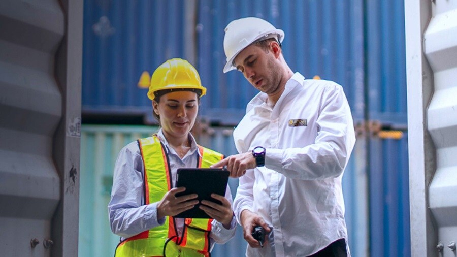 Two logistics workers in safety gear reviewing data on a tablet near stacked shipping containers.
