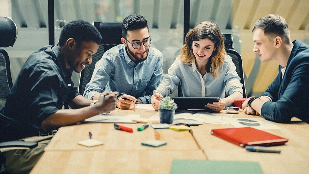 A diverse group of five individuals in a meeting, examining documents on a tablet, with notebooks and pens on the table.