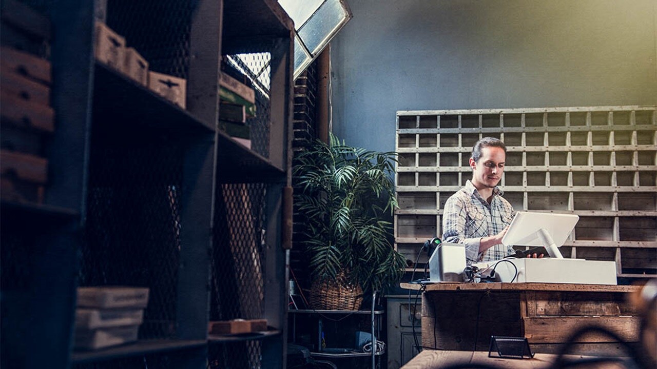 A small business owner working on his computer in front of shelves.