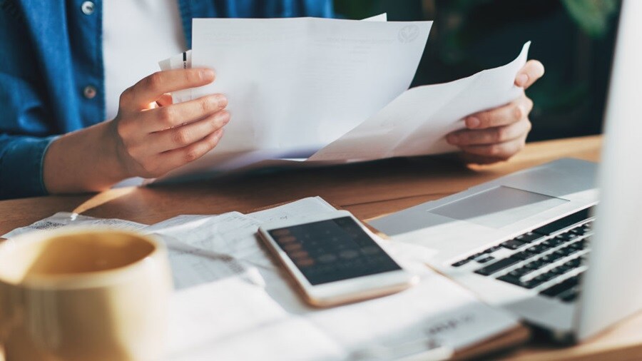 Person reviewing documents at a desk with a laptop, smartphone, and coffee cup.
