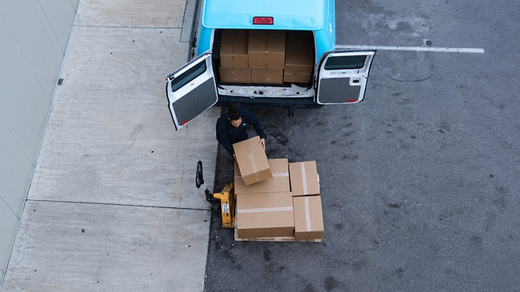An aerial view of a delivery worker loading cardboard boxes from a pallet jack into the back of a light blue van parked on asphalt pavement.