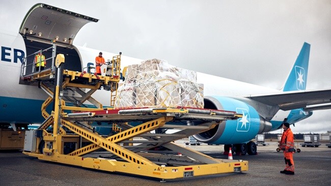 Ground crew load a large wrapped cargo pallet into an open aircraft hold using a yellow hydraulic loader, beneath a cloudy airport sky.