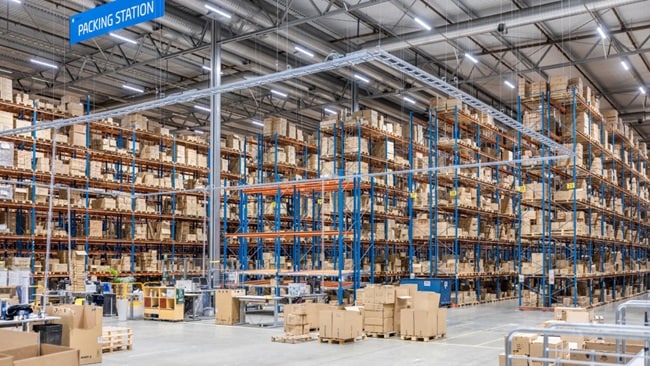 A vast warehouse interior featuring tall blue metal shelving units filled with stacked cardboard boxes and a blue sign labeled Packing Station.