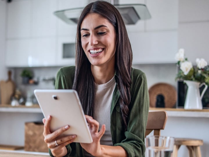 A smiling woman in a green shirt using a tablet at a wooden table in a modern kitchen, with a glass of water nearby.