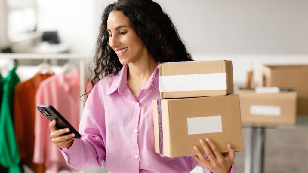 A woman holds a package in one hand while looking at her phone with a focused expression.