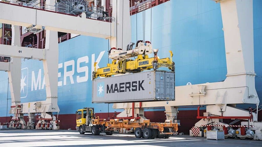 Maersk container being loaded onto a truck by a crane at a port with a large Maersk ship in the background.