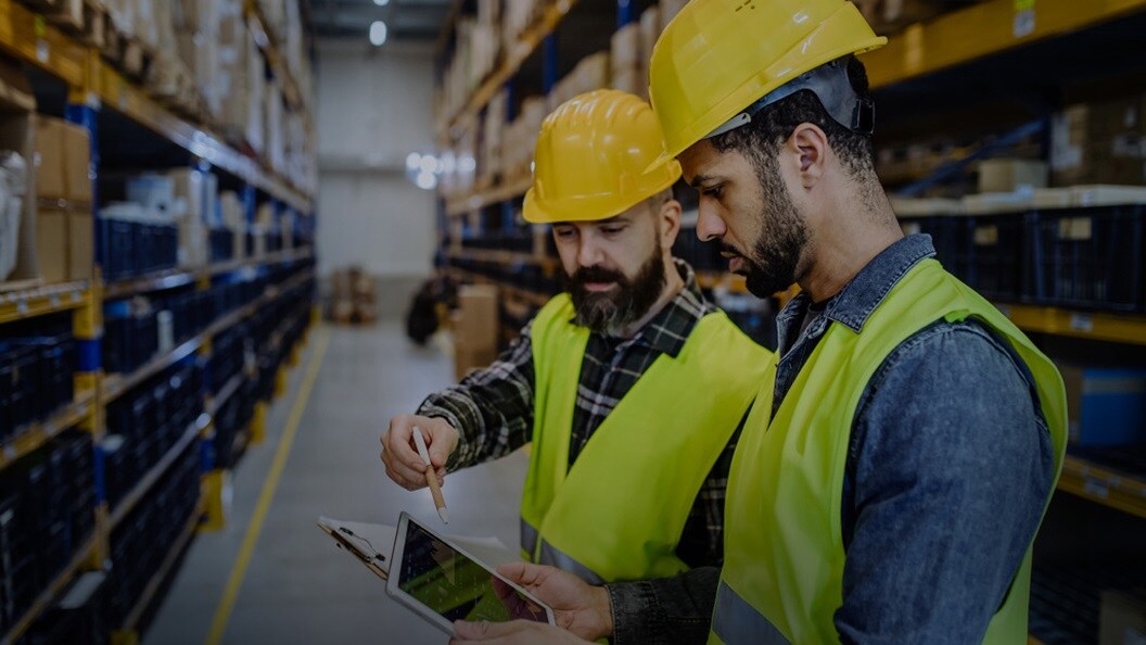 Two persons wearing yellow jackets and yellow hats in a warehouse