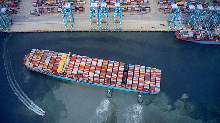 Aerial view of a Maersk container ship being guided by tugboats near a busy port with cranes and stacked containers.