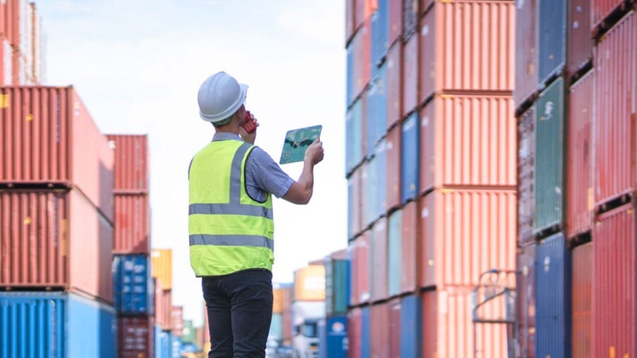 A worker in a hard hat and safety vest inspects stacked shipping containers while using a tablet and phone.