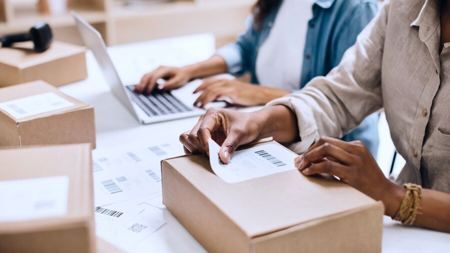 A woman sits at a table, using a laptop while a cardboard box is placed beside her.