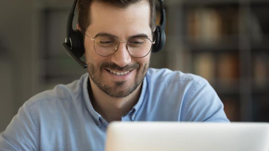 A man wearing glasses and a headset smiles while working on a laptop, conveying a sense of enjoyment and concentration.