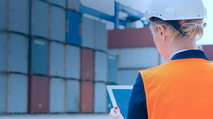 A woman wearing an orange vest and hard hat is holding a tablet, focused on her work in a construction environment.