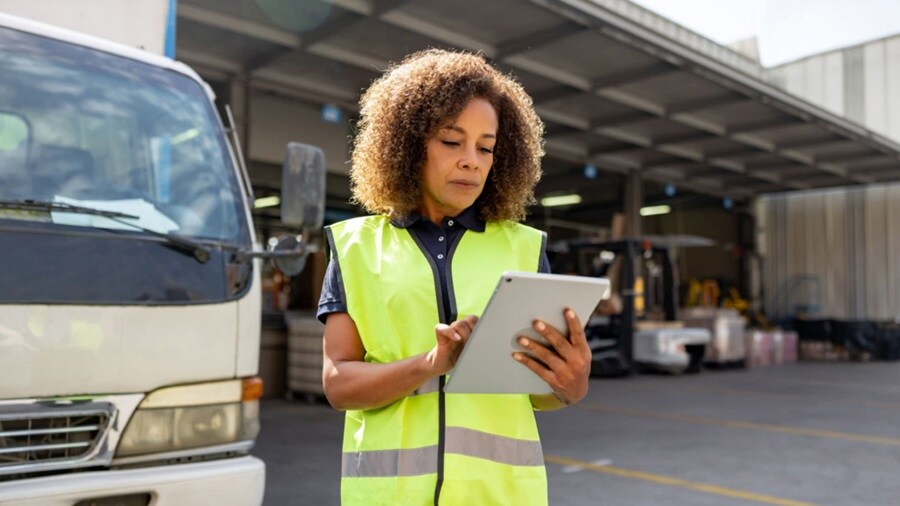 A woman wearing a safety vest holds a tablet computer, focused on her task in a work environment.