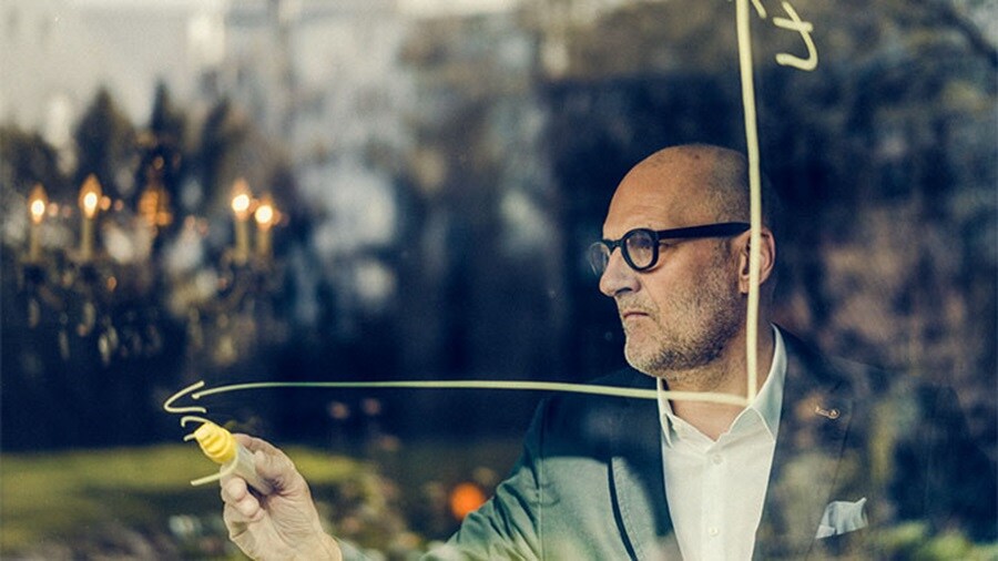 Homem desenhando um gráfico em vidro com um marcador amarelo, visto através de uma janela refletora.