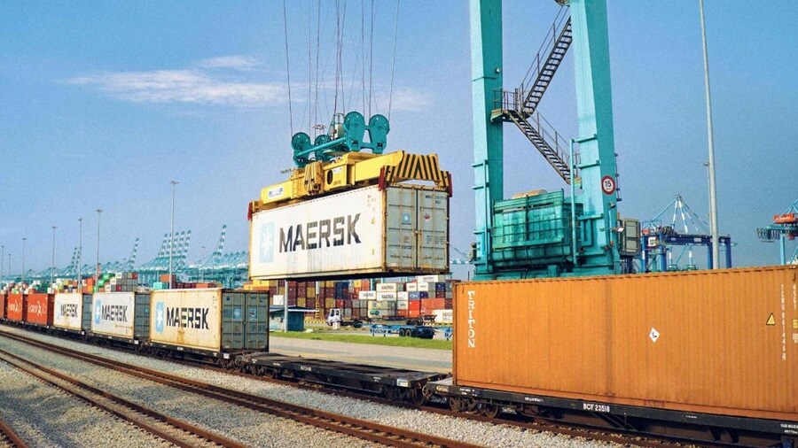 Maersk container being loaded onto a freight train at a busy port terminal.