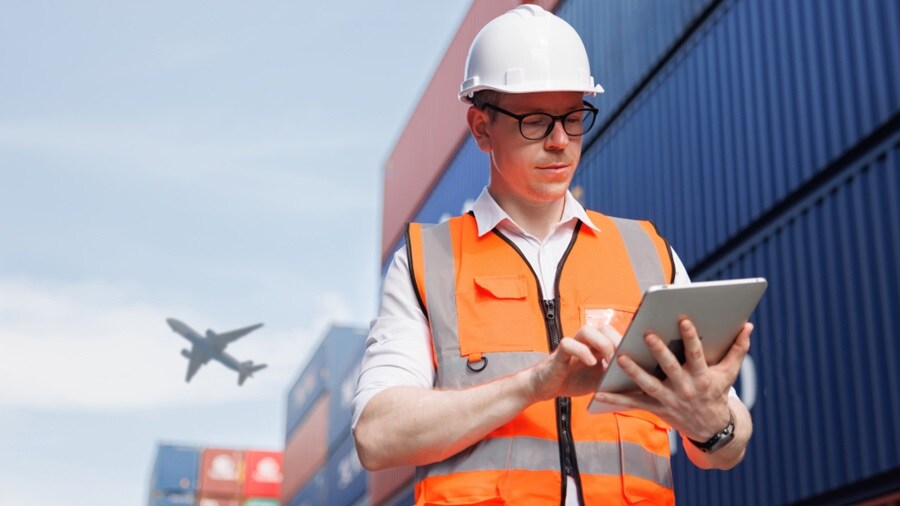 Logistics worker using tablet near cargo containers, with airplane in background.