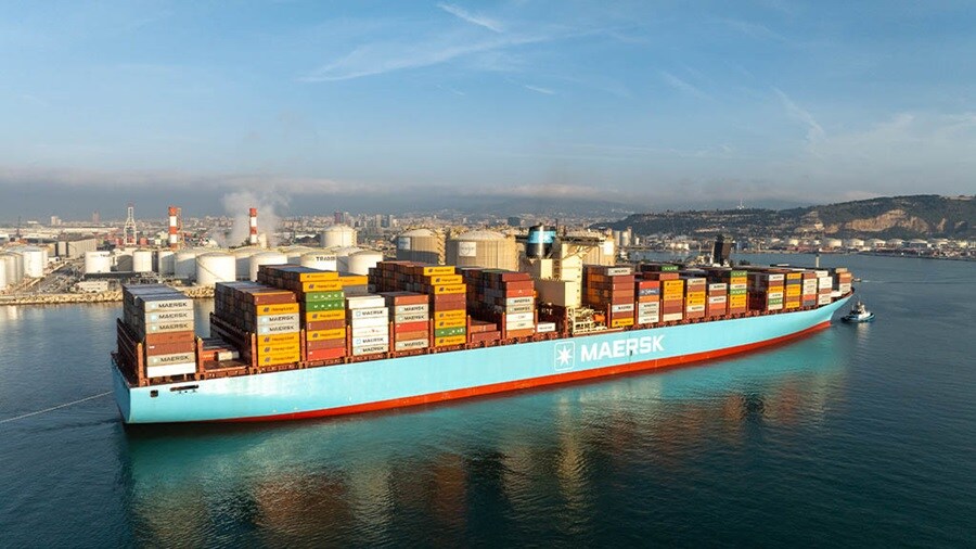 A Maersk container ship sails near an industrial port, stacked high with colorful cargo containers.