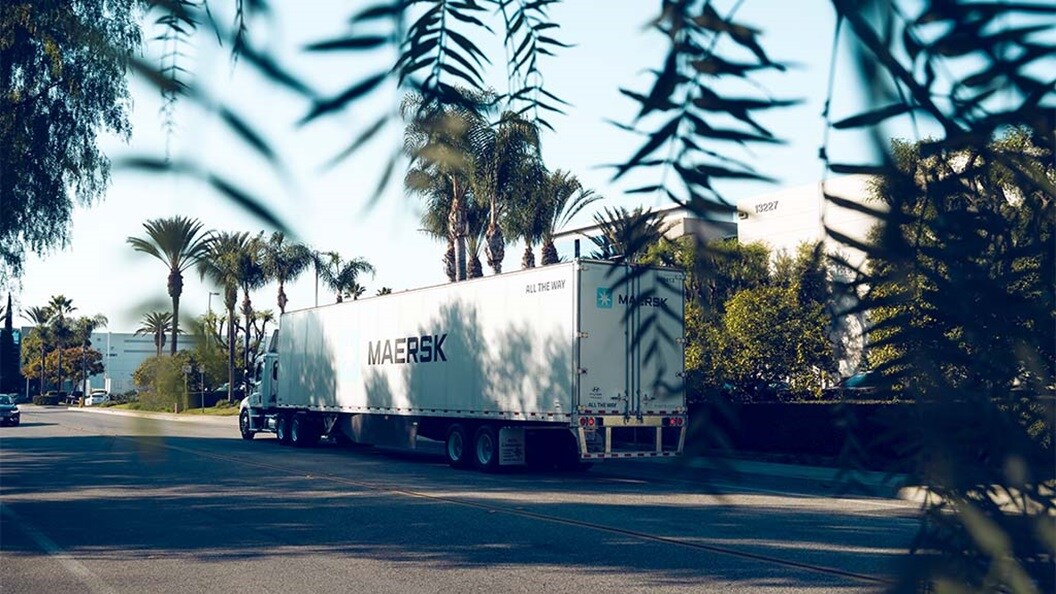A Maersk truck trailer driving through a sunny, palm-lined urban street.