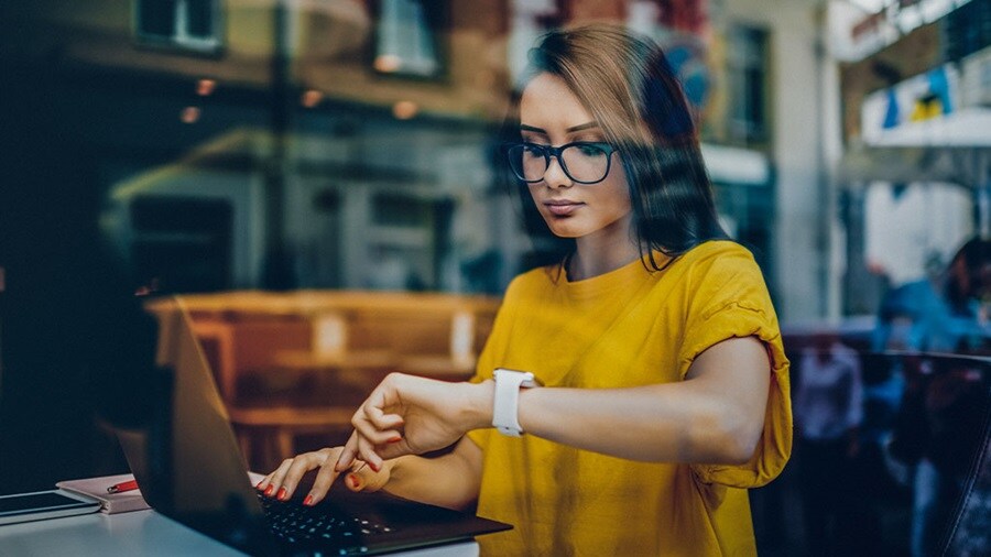 Woman in yellow shirt working on laptop and checking her smartwatch at a cafe.