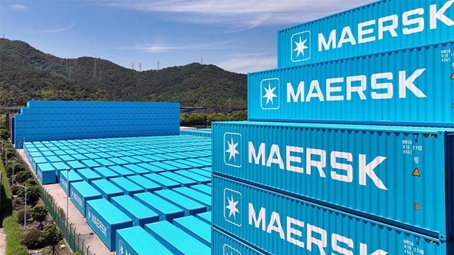 Stacks of blue Maersk shipping containers are arranged in rows at a storage facility, with green hills and a partly cloudy sky in the background.