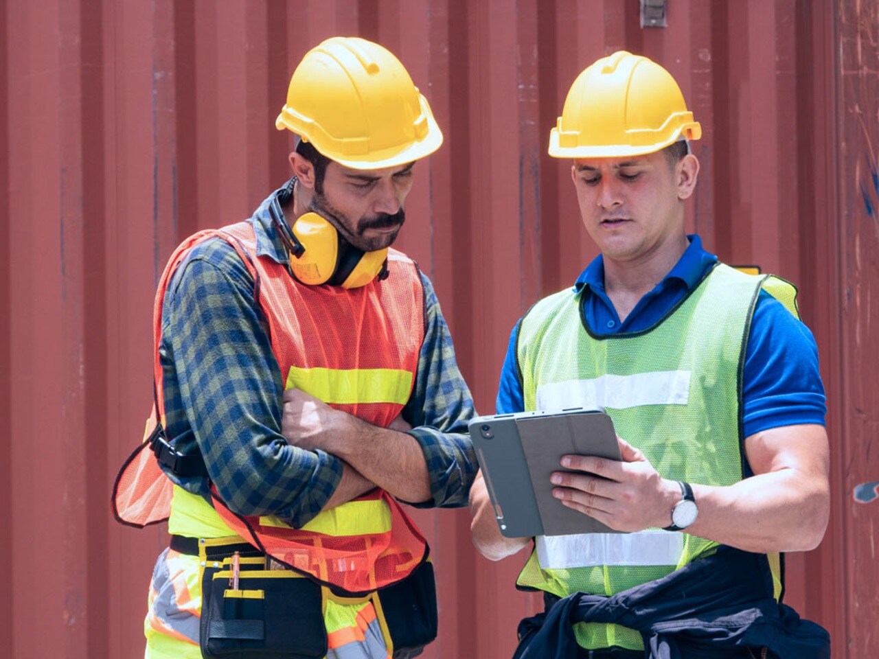 Two workers wearing safety helmets and vests reviewing supply chain operations on a digital tablet at container yard