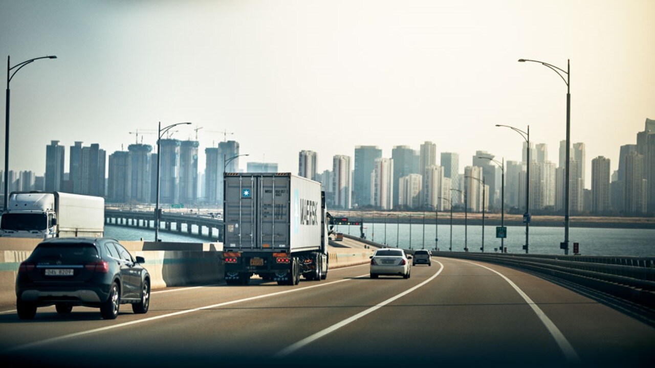 A Maersk container truck drives on a bridge toward a modern city skyline.