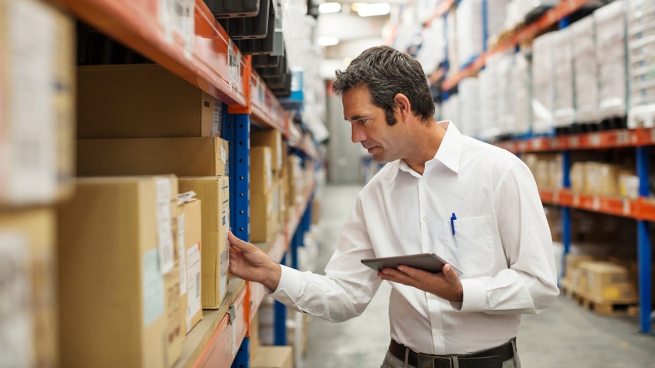 A warehouse worker using a tablet to verify inventory, surrounded by storage shelves and boxes.