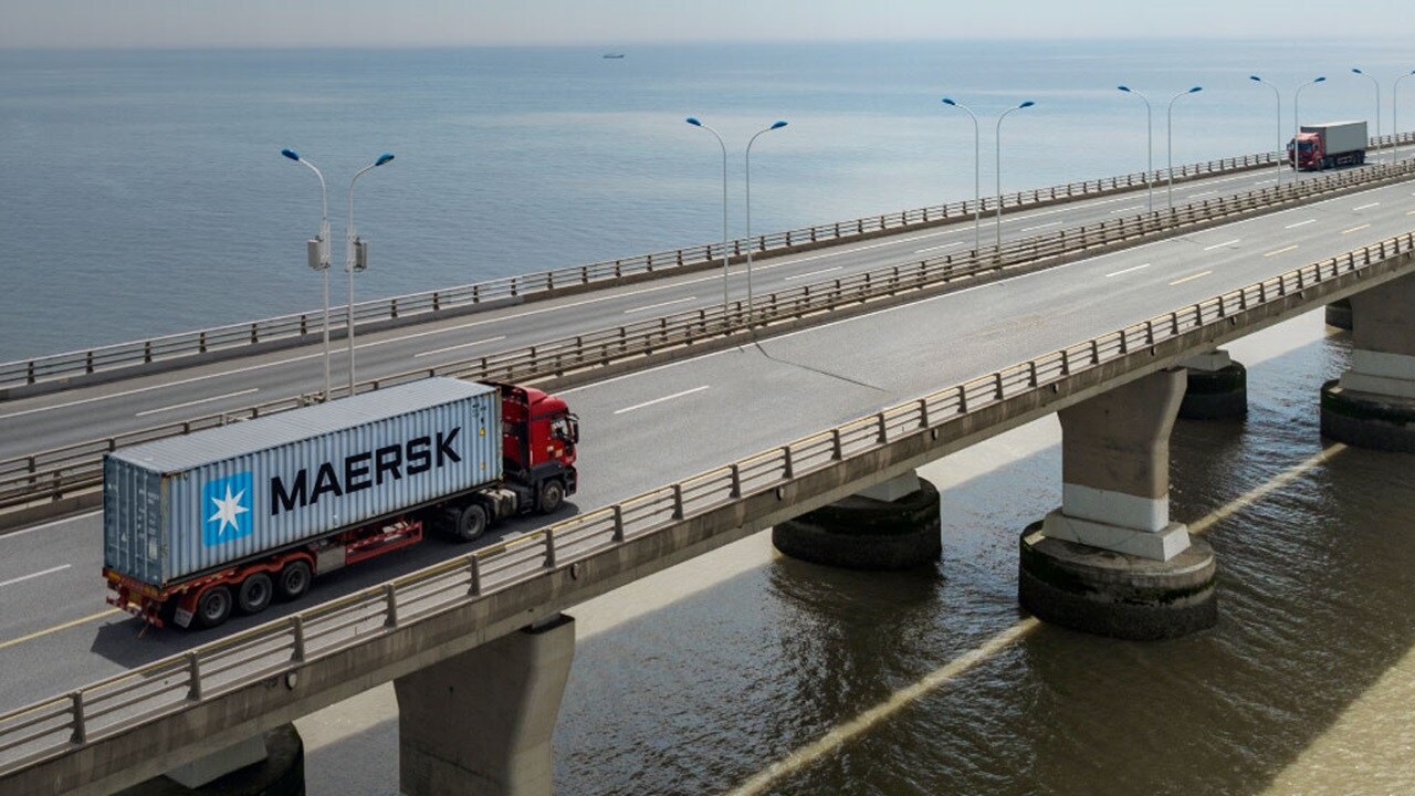 A Maersk container truck driving on a coastal bridge over calm waters, with another truck visible on a parallel road in the background.